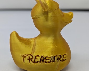 Puede incluir: Una figura de pato dorado con la palabra "TREASURE" grabada en la parte delantera. El pato tiene un pequeño sombrero y está hecho de un material metálico brillante. La figura es un objeto decorativo.