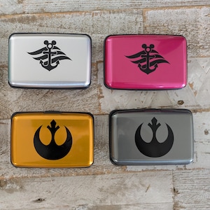May include: Four rectangular card holders in silver, pink, gold, and grey. Each has a black graphic design. The silver and pink holders feature an anchor design with Mickey Mouse ears. The gold and grey holders have a rebel alliance symbol.