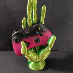 May include: A green skeletal hand holding a pink and black video game controller. The hand is a 3D printed object.