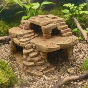 May include: A tan, multi-level stone-like structure with a textured surface resembling stacked rocks. The object features a flat top platform, a central opening, and integrated steps leading to the upper level, designed for use in a terrarium.