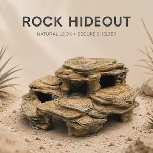 Op de afbeelding: Een beige en bruine rots-schuilplaats met gelaagde, getextureerde oppervlakken en meerdere openingen. De tekst ROCK HIDEOUT NATURAL LOOK • SECURE SHELTER staat boven het object, dat is omringd door zand, kleine stenen en woestijnplanten.