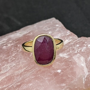 Könnte beinhalten: Ein goldener Ring mit einem großen, ovalen Rubin-Edelstein. Der Rubin ist tiefrot und hat einen facettierten Schliff.