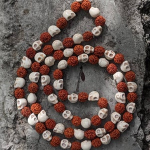 Puede incluir: Un mala hecho de cuentas rojas y blancas, con cuentas en forma de calavera. El mala está sobre un fondo gris.