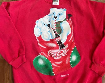 Puede incluir: Sudadera roja de Coca-Cola con un gráfico de dos osos polares bebiendo Coca-Cola de una media. El texto "Coca-Cola" está impreso en la media.