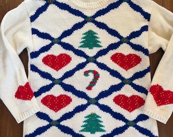 Op de afbeelding: Crèmekleurige gebreide trui met een blauw ruitpatroon. De trui heeft rode harten, groene kerstbomen en een zuurstokontwerp. De mouwen hebben rode hartaccenten. De merknaam "KAREN SCOTT" is zichtbaar op de kraag.