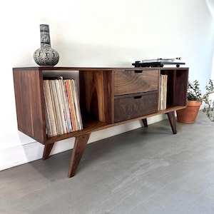 Puede incluir: Un mueble multimedia de madera marrón oscuro con dos cajones apilados en el centro, estantes abiertos a ambos lados para guardar discos de vinilo y cuatro patas inclinadas. La superficie superior sostiene un jarrón estampado y un tocadiscos negro.