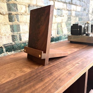Peut inclure: Un support en bois brun fonc&eacute; pour tablette ou t&eacute;l&eacute;phone, compos&eacute; d'un panneau arri&egrave;re vertical et d'une base horizontale reli&eacute;s par un petit rebord avant. Le rebord est fix&eacute; par deux petites goupilles m&eacute;talliques de couleur laiton.