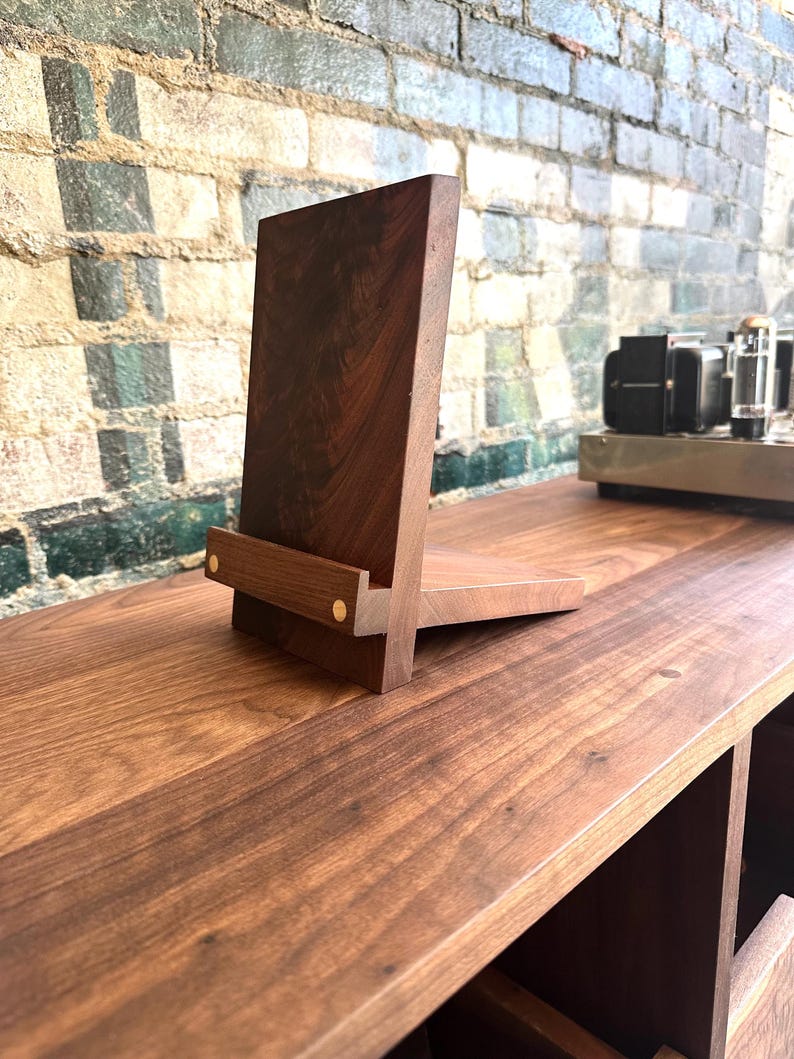 Peut inclure: Un support en bois brun fonc&eacute; pour tablette ou t&eacute;l&eacute;phone, compos&eacute; d'un panneau arri&egrave;re vertical et d'une base horizontale reli&eacute;s par un petit rebord avant. Le rebord est fix&eacute; par deux petites goupilles m&eacute;talliques de couleur laiton.