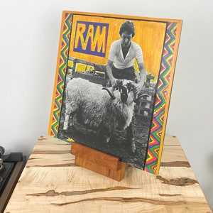 Peut inclure: Une pochette de disque vinyle carr&eacute;e pour l'album RAM pr&eacute;sentant une photographie en noir et blanc d'une personne tenant un b&eacute;lier. La pochette a un fond jaune, le mot RAM en lettres bleues et jaunes, et une bordure en zigzag rouge, bleue et verte.