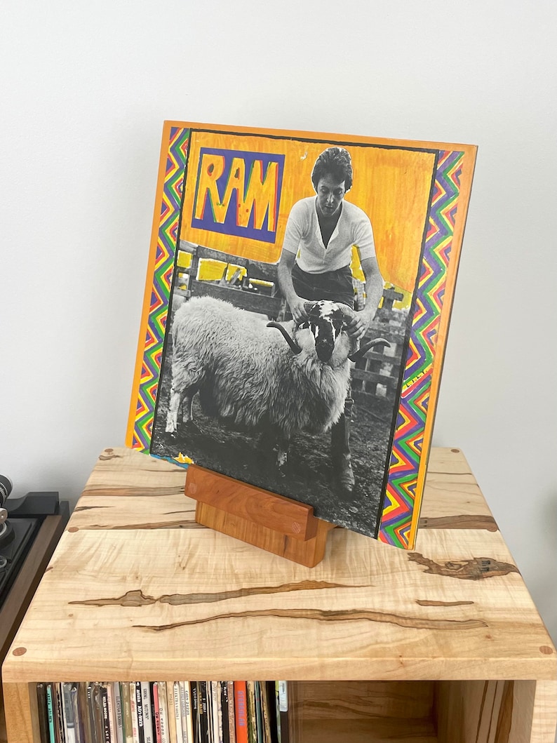 Peut inclure: Une pochette de disque vinyle carr&eacute;e pour l'album RAM pr&eacute;sentant une photographie en noir et blanc d'une personne tenant un b&eacute;lier. La pochette a un fond jaune, le mot RAM en lettres bleues et jaunes, et une bordure en zigzag rouge, bleue et verte.