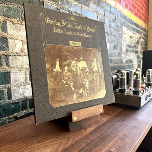Peut inclure: Une pochette de disque vinyle carr&eacute;e pour Crosby, Stills, Nash & Young intitul&eacute;e D&eacute;j&agrave; vu, avec une couverture textur&eacute;e marron fonc&eacute;, des lettres dor&eacute;es et une photographie s&eacute;pia du groupe et d'un chien, pos&eacute;e sur un support en bois.
