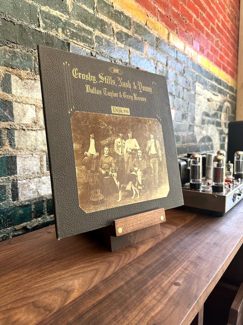 Peut inclure: Une pochette de disque vinyle carr&eacute;e pour Crosby, Stills, Nash & Young intitul&eacute;e D&eacute;j&agrave; vu, avec une couverture textur&eacute;e marron fonc&eacute;, des lettres dor&eacute;es et une photographie s&eacute;pia du groupe et d'un chien, pos&eacute;e sur un support en bois.