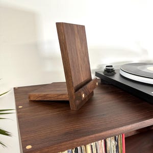 Peut inclure: Un pr&eacute;sentoir en bois brun fonc&eacute; compos&eacute; d'un panneau arri&egrave;re vertical et d'une tablette avant horizontale, dot&eacute; de deux petites incrustations circulaires en bois clair sur la barre de support avant pour maintenir des objets debout.
