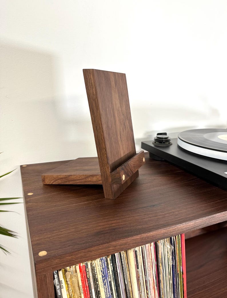 Peut inclure: Un pr&eacute;sentoir en bois brun fonc&eacute; compos&eacute; d'un panneau arri&egrave;re vertical et d'une tablette avant horizontale, dot&eacute; de deux petites incrustations circulaires en bois clair sur la barre de support avant pour maintenir des objets debout.