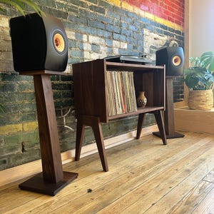 Peut inclure: Un meuble à vinyles en bois foncé avec des pieds inclinés, contenant une collection de disques et un petit vase en céramique. Deux enceintes noires avec des haut-parleurs circulaires dorés sont posées sur des pieds en bois assortis.
