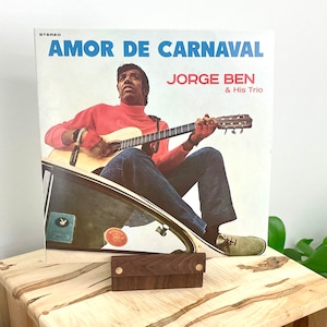 Peut inclure: Une pochette de disque vinyle intitul&eacute;e AMOR DE CARNAVAL par JORGE BEN & His Trio. La pochette montre une personne portant un col roul&eacute; rouge et un jean bleu jouant de la guitare acoustique assise sur le bord d'une voiture. Texte en bleu et rouge.