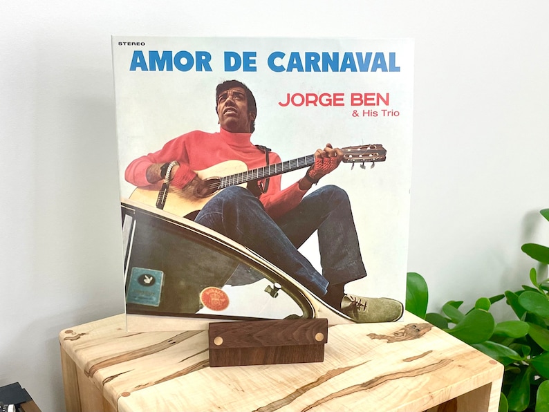 Peut inclure: Une pochette de disque vinyle intitul&eacute;e AMOR DE CARNAVAL par JORGE BEN & His Trio. La pochette montre une personne portant un col roul&eacute; rouge et un jean bleu jouant de la guitare acoustique assise sur le bord d'une voiture. Texte en bleu et rouge.