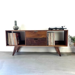 Puede incluir: Una consola de medios de madera de estilo moderno de mediados de siglo con un tocadiscos, discos de vinilo y un jarrón decorativo. La consola tiene dos cajones y espacios de almacenamiento abiertos. La unidad está soportada por patas anguladas y está junto a una planta en maceta.