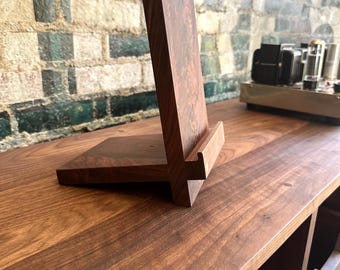 Puede incluir: Un soporte de madera de color marrón oscuro para teléfono que cuenta con un panel trasero vertical con veta de madera visible y una base horizontal con un pequeño borde frontal para sostener un dispositivo móvil. Hecho de madera maciza.
