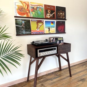 May include: A dark wood record console with a turntable and silver receiver on top, featuring a storage compartment for vinyl records. Two matching wall-mounted shelves above display eight vinyl record covers in a two-row arrangement.