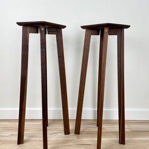 May include: Two dark brown wooden plant stands with a square top and four legs. The stands are made of solid wood and have a simple, modern design.