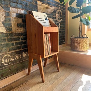 Può includere: Un supporto in legno marrone chiaro per dischi in vinile con gambe inclinate. L'unità presenta una sezione superiore aperta per esporre la copertina di un disco e un ripiano rettangolare inferiore riempito con una collezione di dischi in vinile.