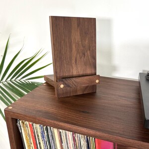 Peut inclure: Un pr&eacute;sentoir en bois brun fonc&eacute; compos&eacute; d'un panneau arri&egrave;re vertical et d'un rebord avant horizontal. Le support pr&eacute;sente deux petits accents circulaires en bois clair sur le rebord avant, mettant en valeur une texture de grain naturel.