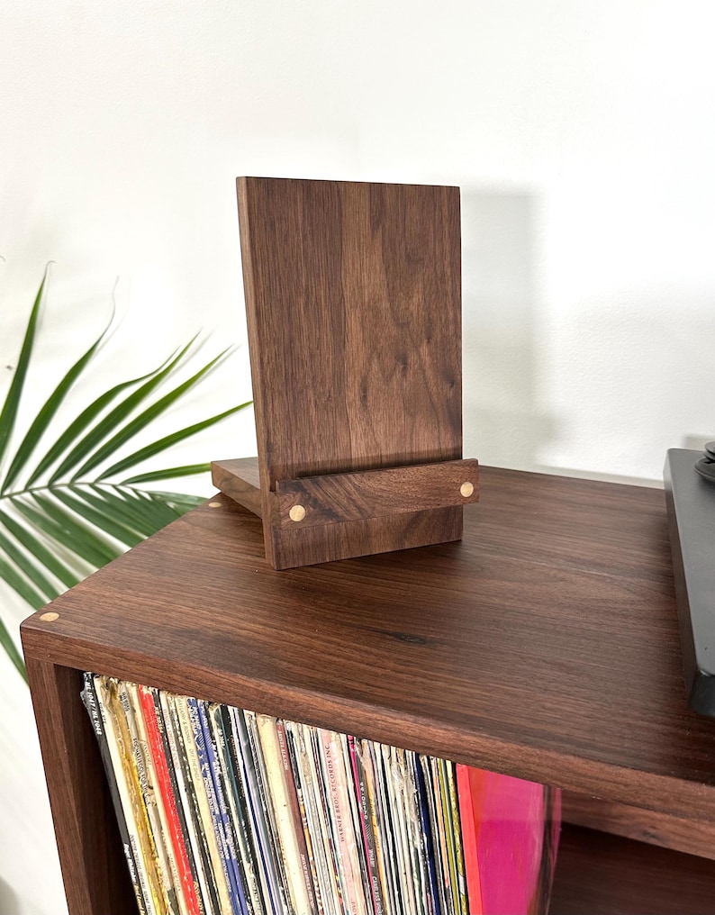 Peut inclure: Un pr&eacute;sentoir en bois brun fonc&eacute; compos&eacute; d'un panneau arri&egrave;re vertical et d'un rebord avant horizontal. Le support pr&eacute;sente deux petits accents circulaires en bois clair sur le rebord avant, mettant en valeur une texture de grain naturel.