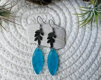 May include: Teal blue glass teardrop earrings with silver leaf accents. The earrings are hanging from silver hooks.