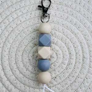 May include: A beige and grey silicone beaded keychain with a silver metal clip. The beads are round and geometric shapes.