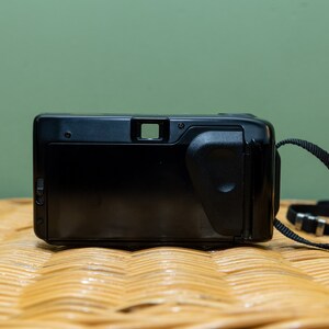 Pu&ograve; includere: Fotocamera a pellicola point-and-shoot nera con cinturino nero. La fotocamera ha una forma rettangolare con mirino e impugnatura strutturata. La fotocamera &egrave; appoggiata su una superficie intrecciata con sfondo verde.