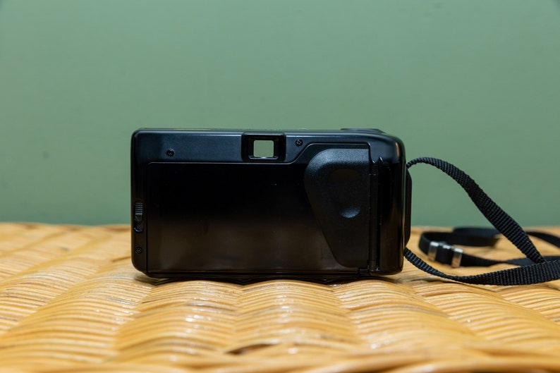 Pu&ograve; includere: Fotocamera a pellicola point-and-shoot nera con cinturino nero. La fotocamera ha una forma rettangolare con mirino e impugnatura strutturata. La fotocamera &egrave; appoggiata su una superficie intrecciata con sfondo verde.