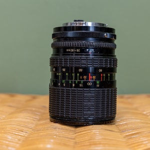 May include: A black camera lens with a silver ring around the front. The lens has a depth of field scale marked in feet and meters, with a range of 28 to 80 mm.