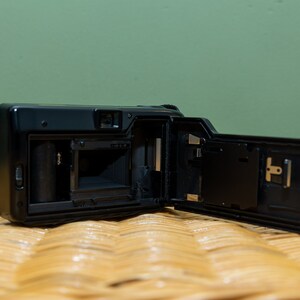 Pu&ograve; includere: Fotocamera a pellicola nera con il retro aperto, che rivela i componenti interni. La fotocamera &egrave; posizionata su una superficie tessuta, con uno sfondo verde. Il retro aperto mostra il vano pellicola e altre parti meccaniche.