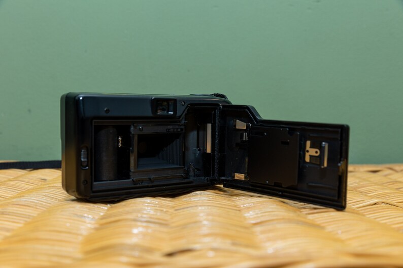 Pu&ograve; includere: Fotocamera a pellicola nera con il retro aperto, che rivela i componenti interni. La fotocamera &egrave; posizionata su una superficie tessuta, con uno sfondo verde. Il retro aperto mostra il vano pellicola e altre parti meccaniche.