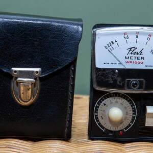 May include: A black leather case with a silver clasp next to a black flash meter with a white face. The flash meter has a dial with numbers and a needle. The text on the face reads "Flash Meter WP1000".