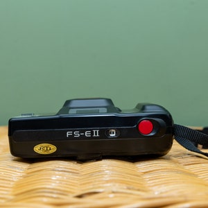 Pu&ograve; includere: Fotocamera FS-EII nera con un pulsante rosso e un cinturino nero. La fotocamera ha un adesivo dorato con testo. &Egrave; su una superficie tessuta con sfondo verde.