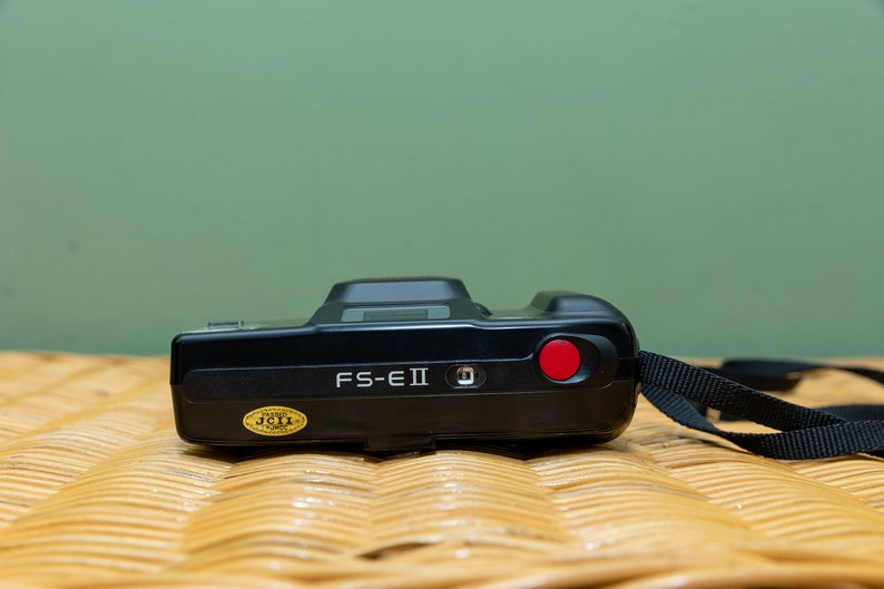 Pu&ograve; includere: Fotocamera FS-EII nera con un pulsante rosso e un cinturino nero. La fotocamera ha un adesivo dorato con testo. &Egrave; su una superficie tessuta con sfondo verde.