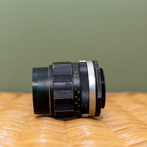 Puede incluir: Un objetivo de cámara negro con un anillo plateado y marcas en verde. El objetivo tiene una escala de profundidad de campo con números del 4 al 100.