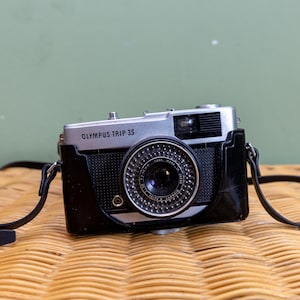 Op de afbeelding: Een zilveren en zwarte Olympus Trip 35 filmcamera met een zwarte leren riem. De camera ligt op een geweven rieten oppervlak.