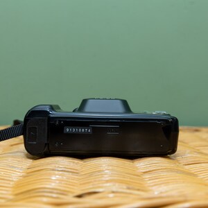 Pu&ograve; includere: Una fotocamera digitale nera con un cinturino nero. La fotocamera ha una piccola forma rettangolare con un numero di serie stampato sul fondo. Lo sfondo &egrave; di colore verde chiaro.