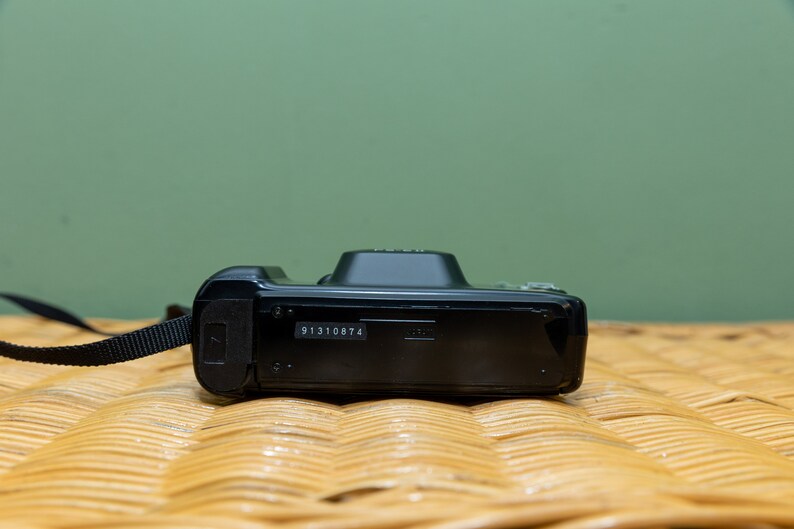 Pu&ograve; includere: Una fotocamera digitale nera con un cinturino nero. La fotocamera ha una piccola forma rettangolare con un numero di serie stampato sul fondo. Lo sfondo &egrave; di colore verde chiaro.