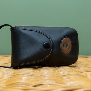 Pu&ograve; includere: Custodia per fotocamera in pelle nera con un accento circolare marrone con il marchio "Minolta". La custodia ha una chiusura a scatto e un cinturino nero. La custodia &egrave; appoggiata su una superficie tessuta.