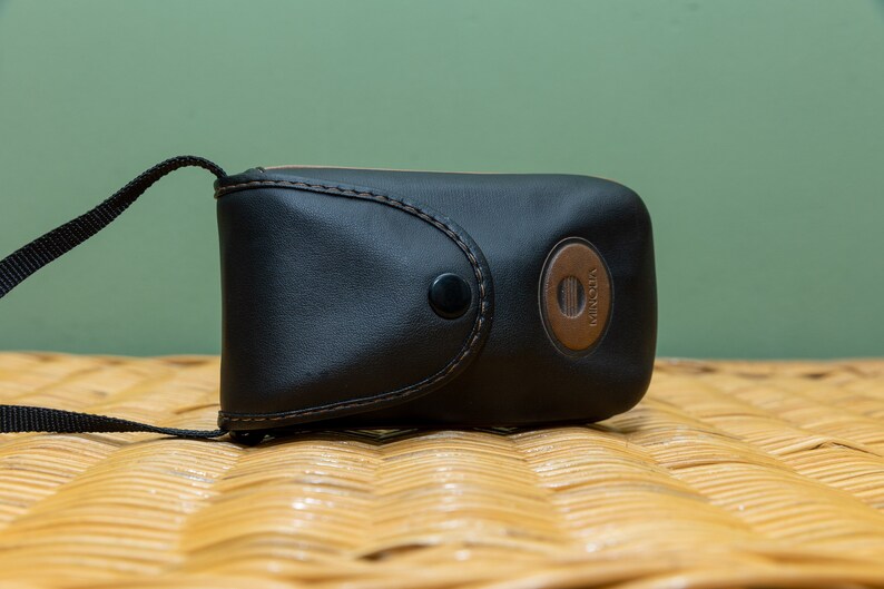 Pu&ograve; includere: Custodia per fotocamera in pelle nera con un accento circolare marrone con il marchio "Minolta". La custodia ha una chiusura a scatto e un cinturino nero. La custodia &egrave; appoggiata su una superficie tessuta.
