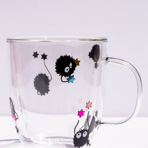 May include: A clear glass mug with a black handle features a whimsical design of black fuzzy creatures with large eyes and colorful stars.