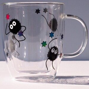 May include: A clear glass mug with a double wall design features black and white illustrations of fuzzy creatures with large eyes and stars. The creatures are depicted with long arms and legs, and the stars are in various colors.