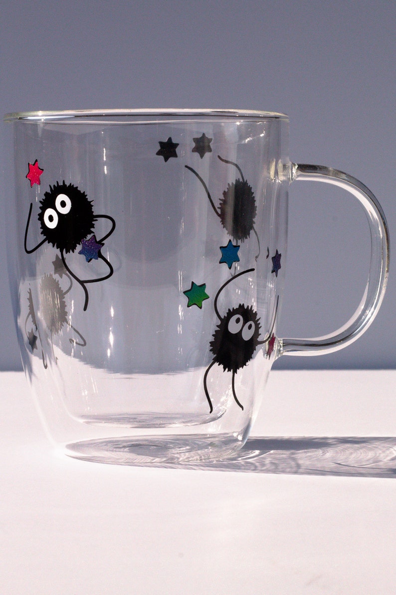 May include: A clear glass mug with a double wall design features black and white illustrations of fuzzy creatures with large eyes and stars. The creatures are depicted with long arms and legs, and the stars are in various colors.