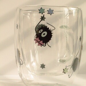 May include: A clear glass double-walled mug with a black and white cartoon character holding a pink star. The character is surrounded by blue and green stars.