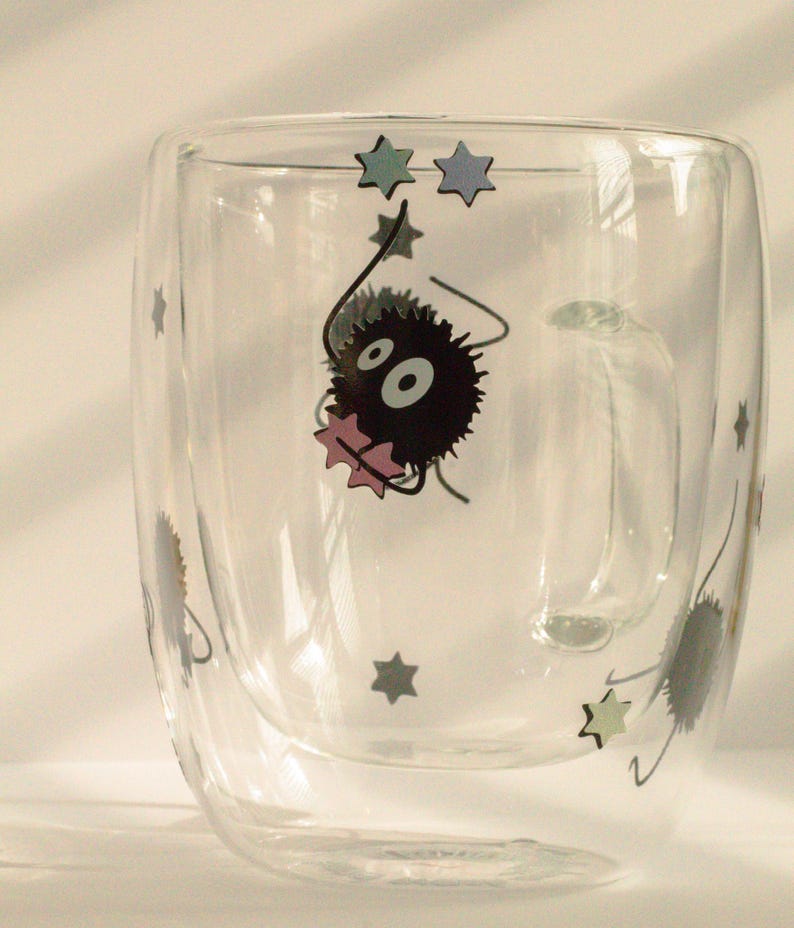May include: A clear glass double-walled mug with a black and white cartoon character holding a pink star. The character is surrounded by blue and green stars.