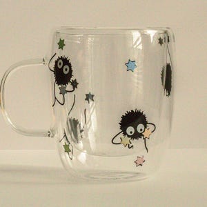 May include: A clear glass mug with a double wall design features black and white illustrations of fuzzy creatures with large eyes and stars. The mug has a handle.